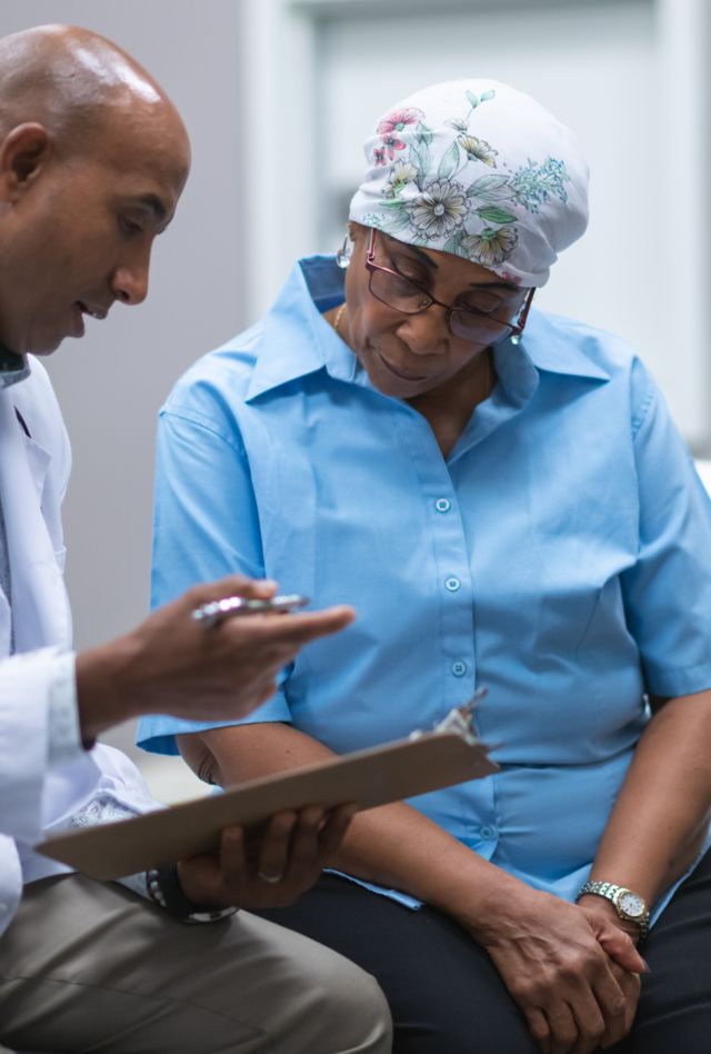 A black senior woman with cancer is at a medical consultation. She is wearing a scarf on her head to hide her hair loss. The doctor is a black man. The patient and doctor are sitting next to each other on an examination table in a medical clinic. The doctor is asking the patient questions and taking notes on a clipboard.