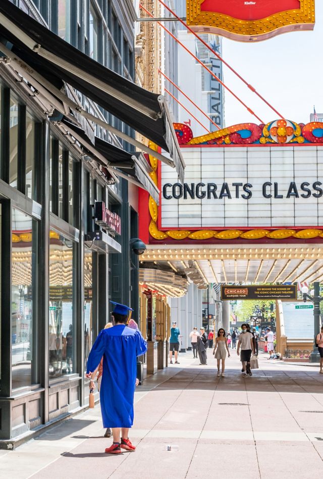 Chicago, IL / USA - May 24,2020: A young man in cap and gown walks under a sign downtown at the Chicago Theater, saying "Congrats Class of 2020."