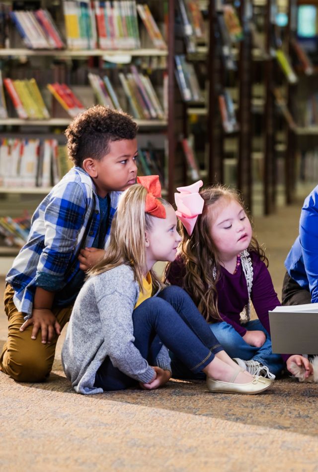 A multi-ethnic group of six boys and girls sitting on the floor of a library, reading with a therapy dog, a goldendoodle. The dog handler is a mature woman in her 50s who is smiling holding a book and reading aloud. The girl 3rd from the left has down syndrome.