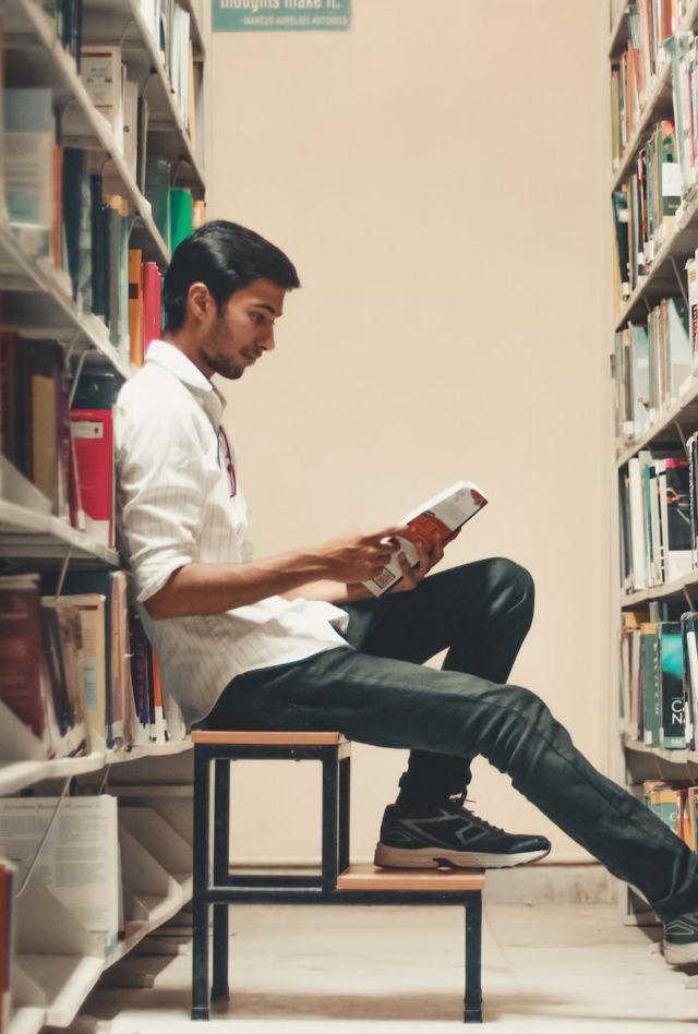 A person sits on a stool reading a book between two library shelves