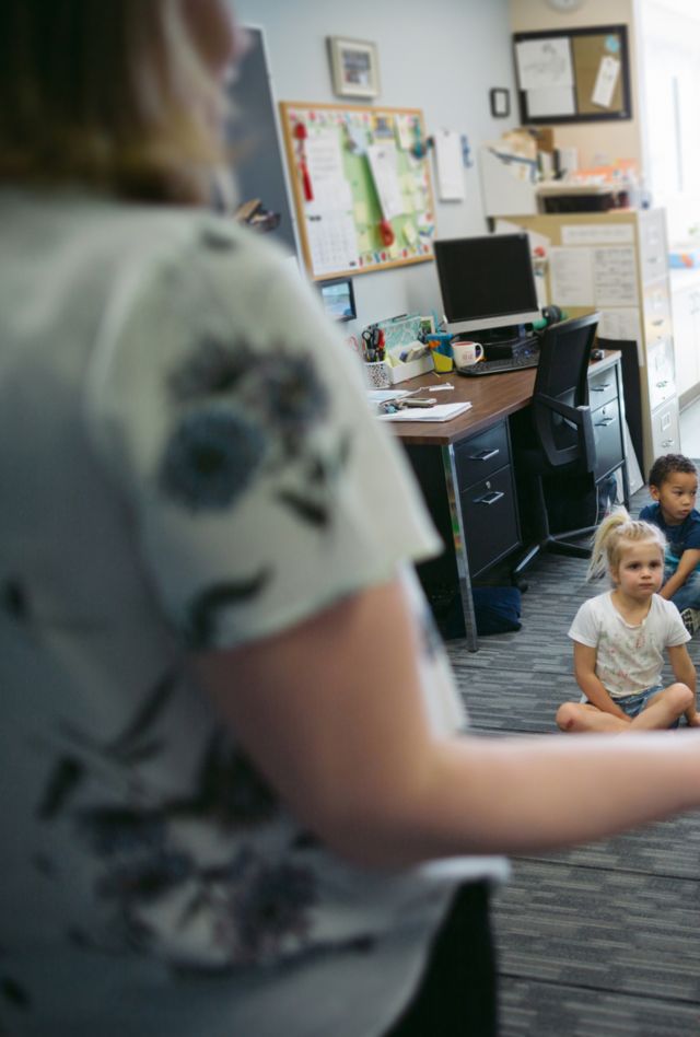 A group of young children sit on the ground in a classroom listening to their teacher