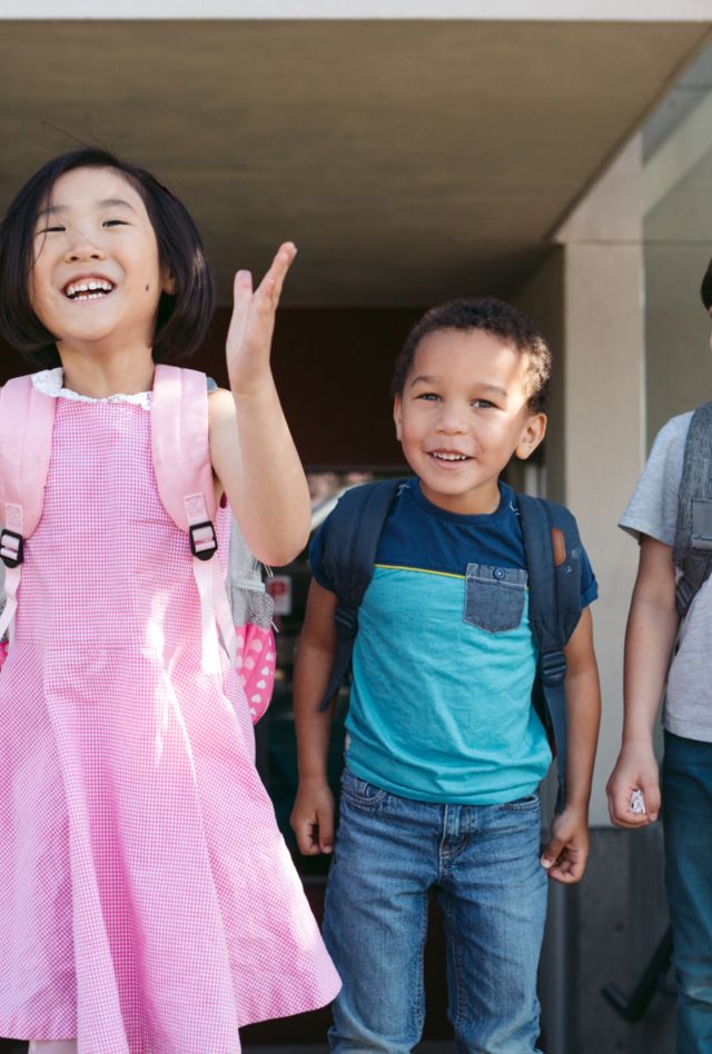 Energetic preschool or kindergarten kids acting goofy outside the school while wearing backpacks.