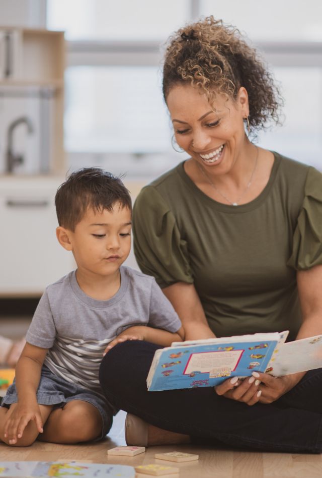 A preschool boy of Hispanic ethnicity sits next to his teacher as she is reading him a book on the classroom floor.