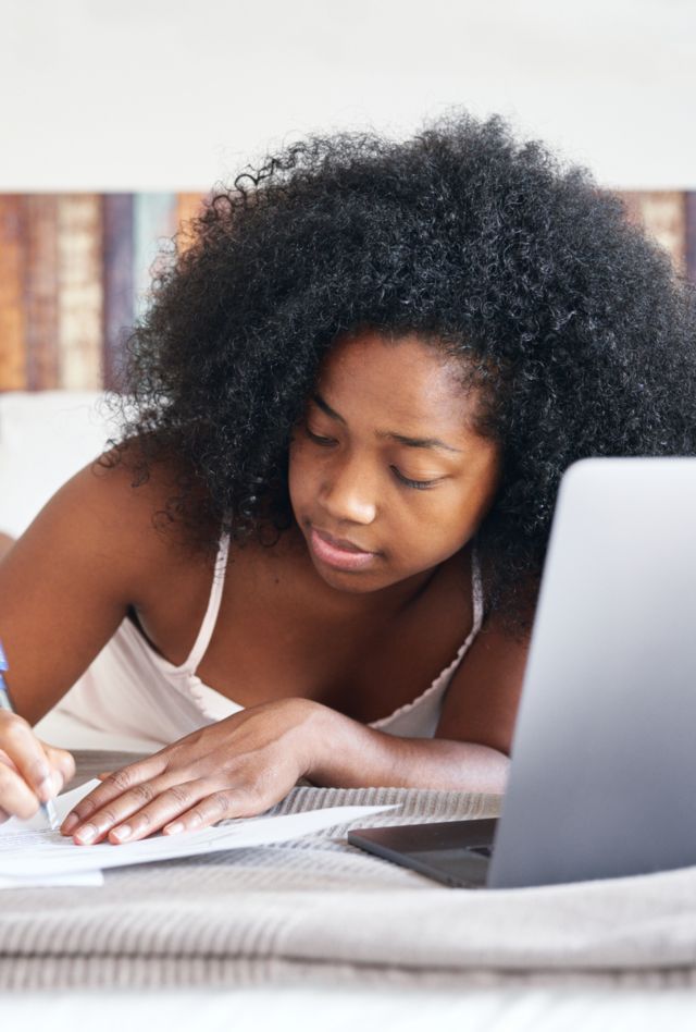 Stock photo of woman with dark frizzy hair in top lying on bed and taking notes with a pen with her laptop in front of her. Working or studying from home during pandemic.