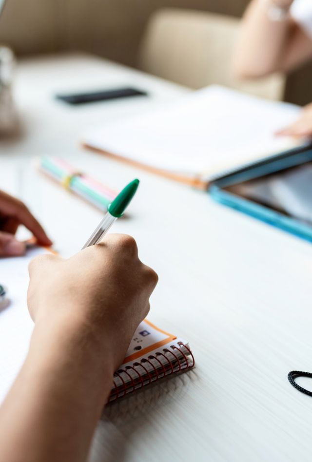 Stock photo of hands of students writing in notebook, using tablet and typing on laptop at one desk. Crop young people studying together and doing homework. Concept of education