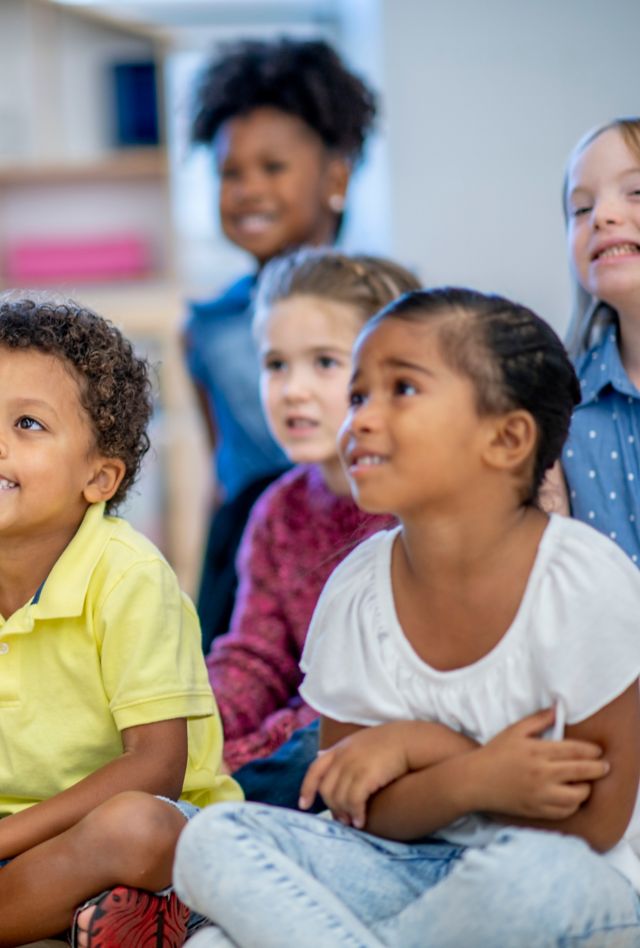 A multi ethnic group of elementary school children are seated together on a classroom floor. They are looking at their teacher and listening intently as their teacher reads them a story from a book.