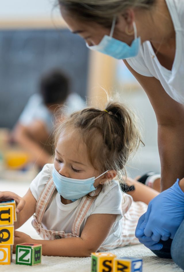 3 year old girl playing at daycare while wearing a protective face mask to protect from the transfer of germs during phase 2 of reopening during COVID-19.