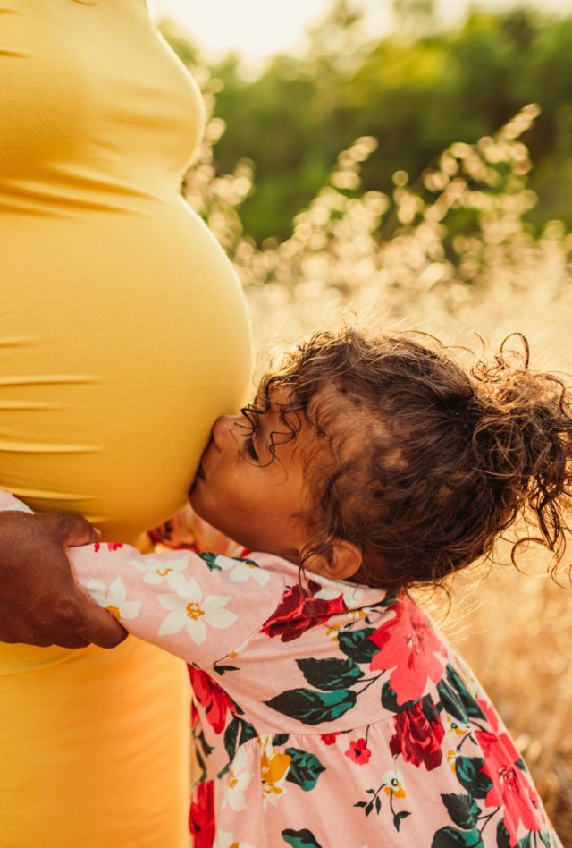 A little girl in a flower dress kisses her mother's baby bump. They are on-location at sunset.