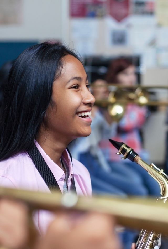 Cheerful Mixed Ethnicity Asian teenage girl sitting with saxophone while looking away. Happy female student is learning woodwind instrument in classroom. She is with friends at high school.