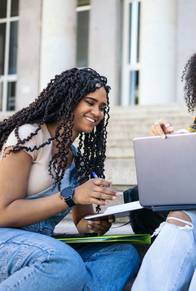 Young students in casual clothes sitting on stairs outdoors at university campus and talking while working on laptop on class project