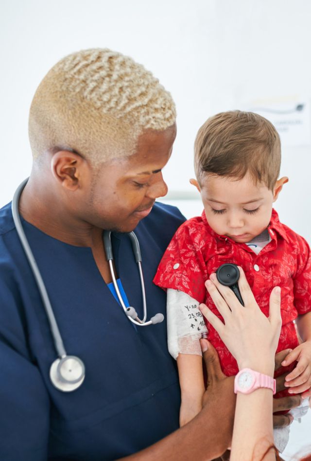 A health provider holds a young child while another provider holds a stethoscope to his chest