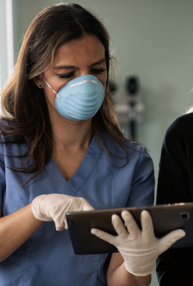 Female patient wearing medical mask in exam room at clinic.  Doctor and nurse also wearing masks to protect from virus or disease.