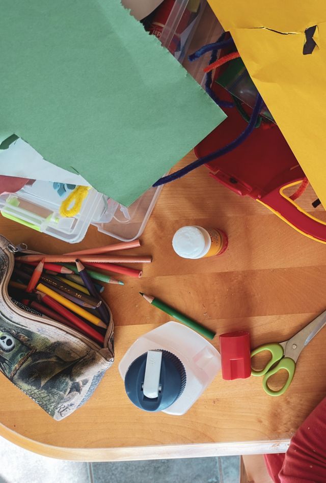 A child draws with a crayon on a surface covered in art supplies and paper