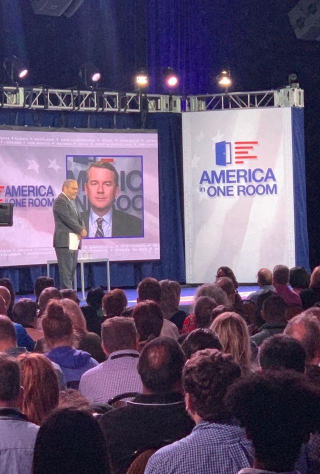 A man stands on a stage in front of a crowd. Behind him is a screen with the America in on room logo. A woman in the crowd is standing up behind a microphone. 