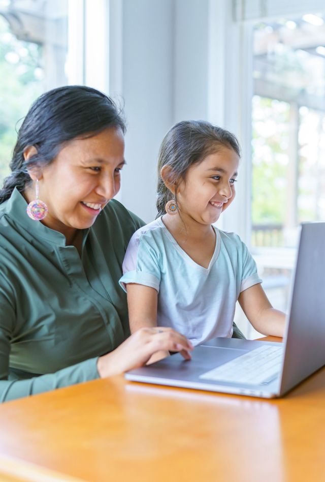 A beautiful Native American mother helps her young daughter with a school assignment online while she studies at home during the COVID-19 pandemic. The woman's adolescent son is sitting at the table working on a school assignment.