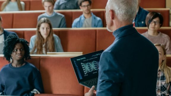 Large group of high school students attending a lecture in the classroom while male teacher is assisting one of them