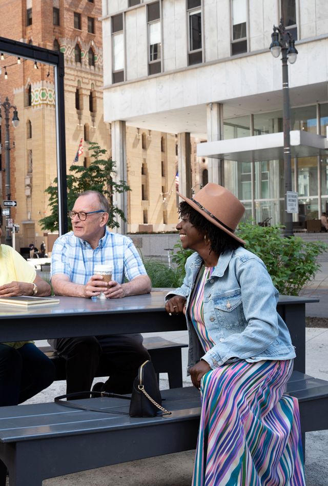 Three people of different races, genders, and ages sitting around a picnic table near large buildings