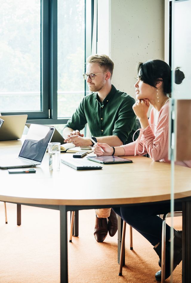 A focused team sits at a round table, deeply engaged in a business meeting with notes and digital devices at hand, in a bright office space.