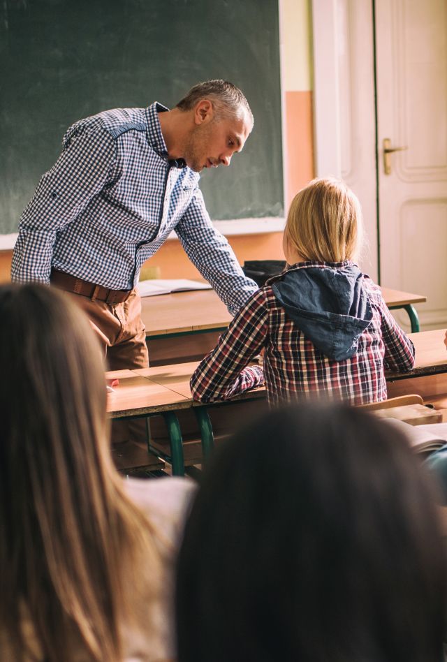 Large group of high school students attending a lecture in the classroom while male teacher is assisting one of them.