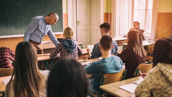 Large group of high school students attending a lecture in the classroom while male teacher is assisting one of them