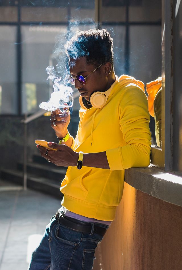 A young black adult male dressed in a yellow sweatshirt and blue pants is smoking leaning against a wall while looking at his cell phone screen.