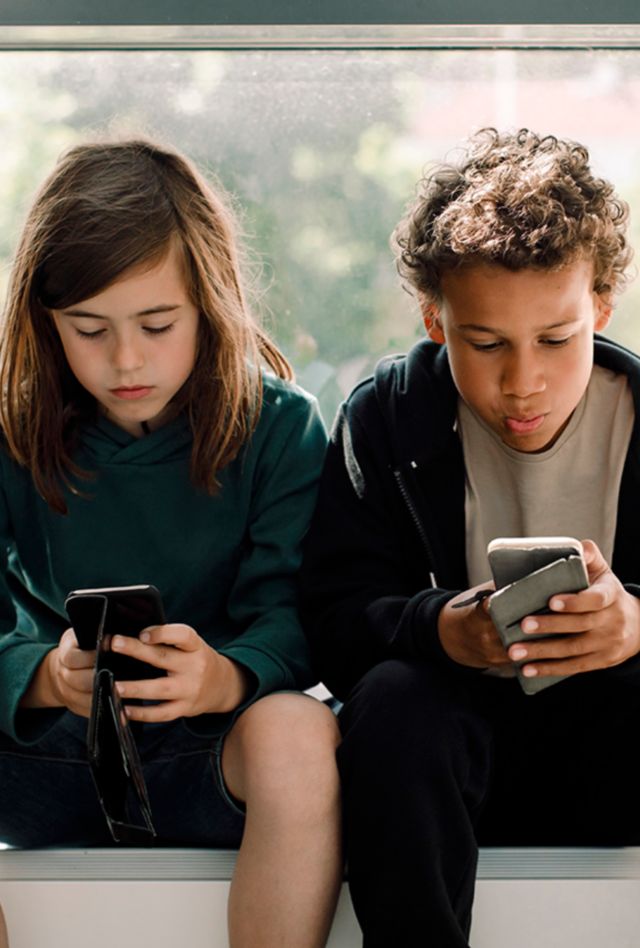 To children sitting in front of a large window looking at cell phones