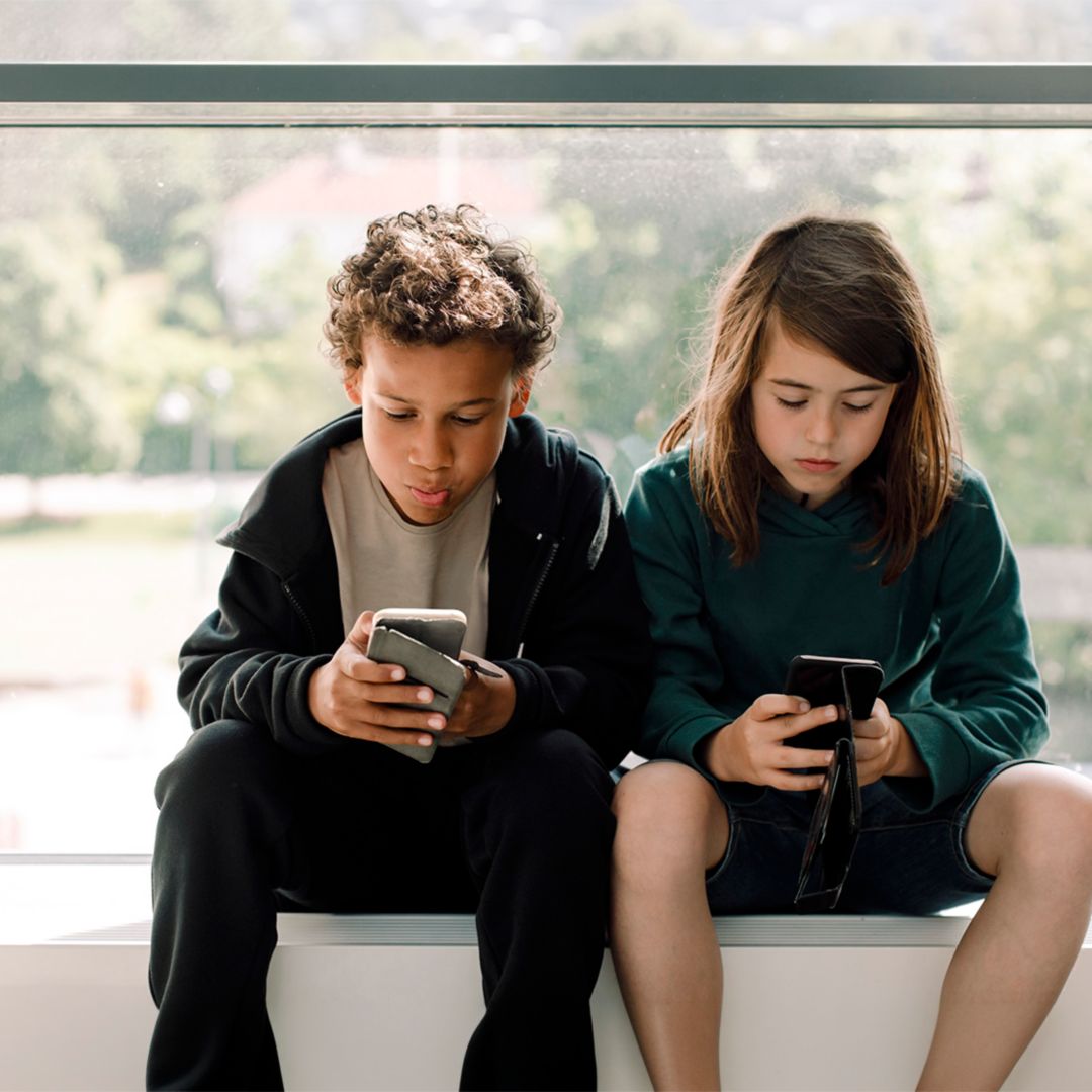Two children sitting in front of a large window looking at cell phones