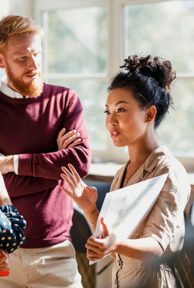 Colleagues standing in a small group discussing something. One of the women is holding documents and gesturing with her hands as the others watch and listen.