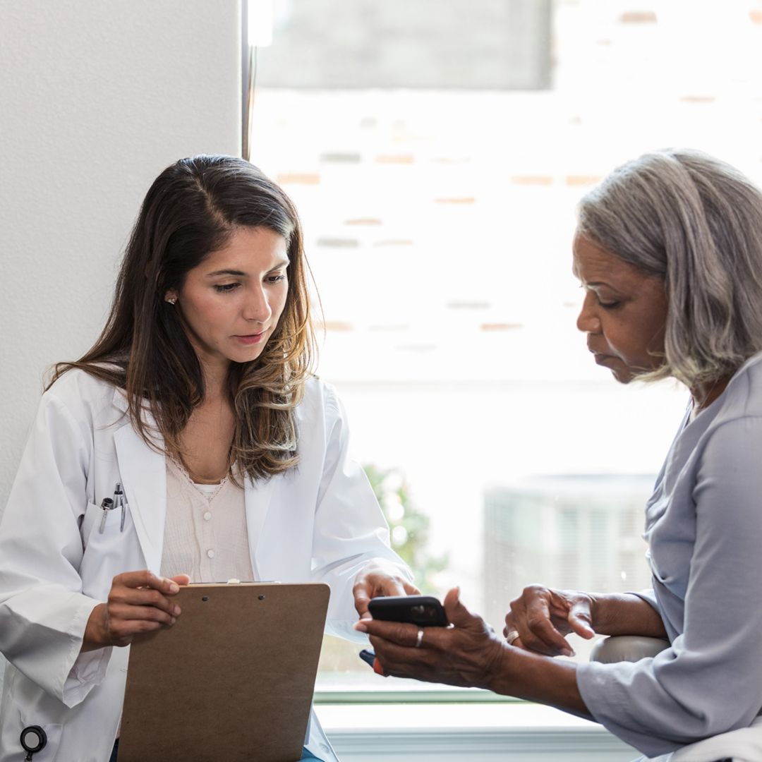 A female doctor in a lab coat shows a clipboard to a patient, who is looking at her smartphone in a medical exam room.