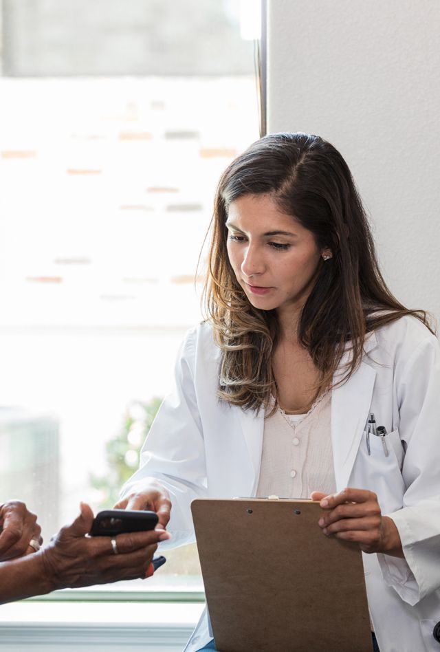A female doctor in a lab coat shows a clipboard to a patient, who is looking at her smartphone in a medical exam room.