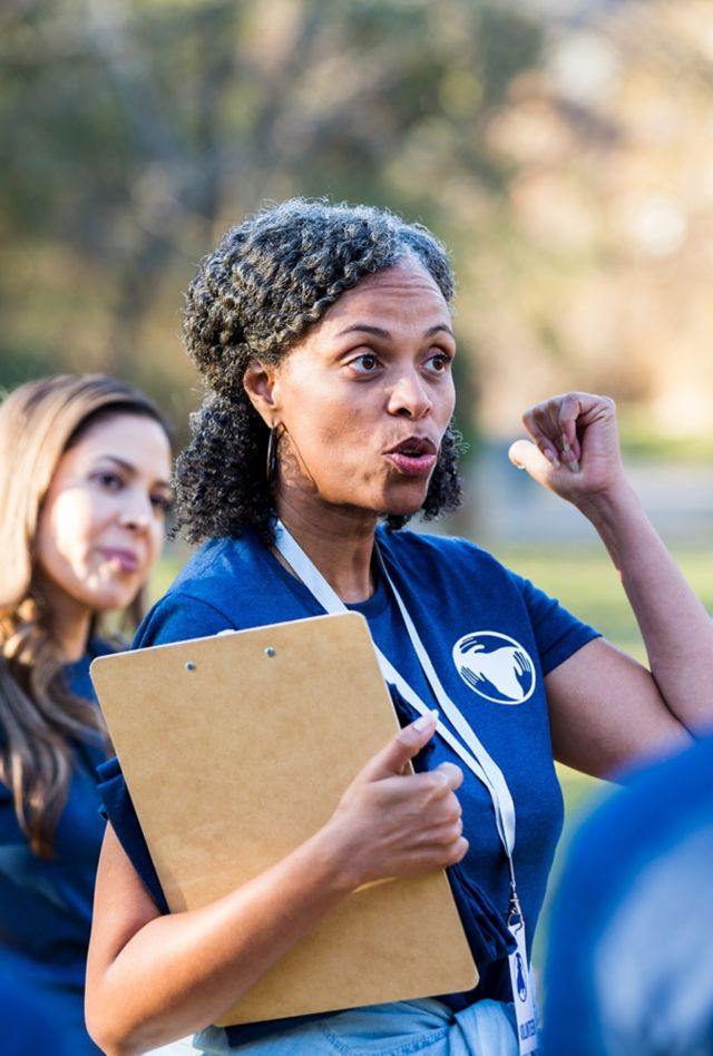 A woman holding a clipboard talks to a group of people who are gathered around her. They are all wearing matching blue shirts.