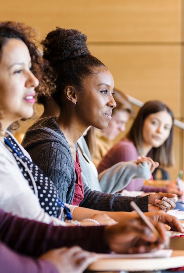 A diverse group of college students are seated in a row in the lecture hall.  Most are engaged in listening to the professor.