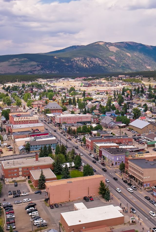Aerial photograph of historic downtown Leadville, Colorado, looking along Harrison Avenue with its rows of brick commercial buildings, church steeples, and surrounding residential neighborhoods. The Mosquito Range rises in the background under a partly cloudy sky, with dense pine forests blanketing the slopes. As the highest incorporated city in North America at over 10,000 feet, Leadville's compact downtown grid sits in a broad mountain valley surrounded by Colorado's tallest peaks.