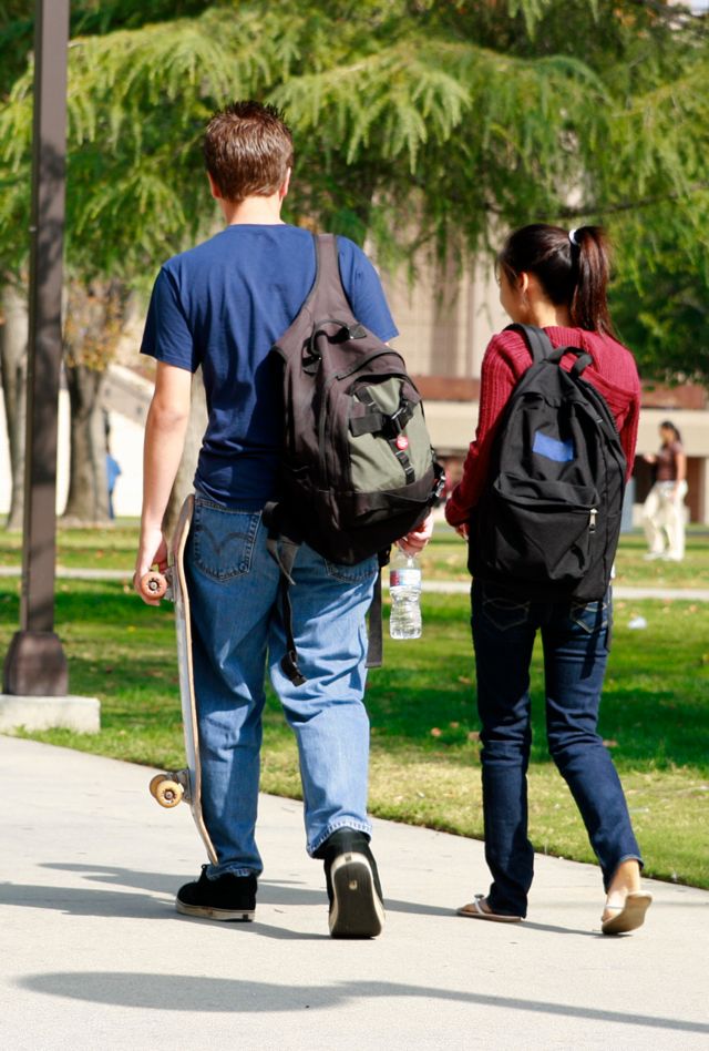 Rearview to young people wearing backpacks walking on college campus sidewalk