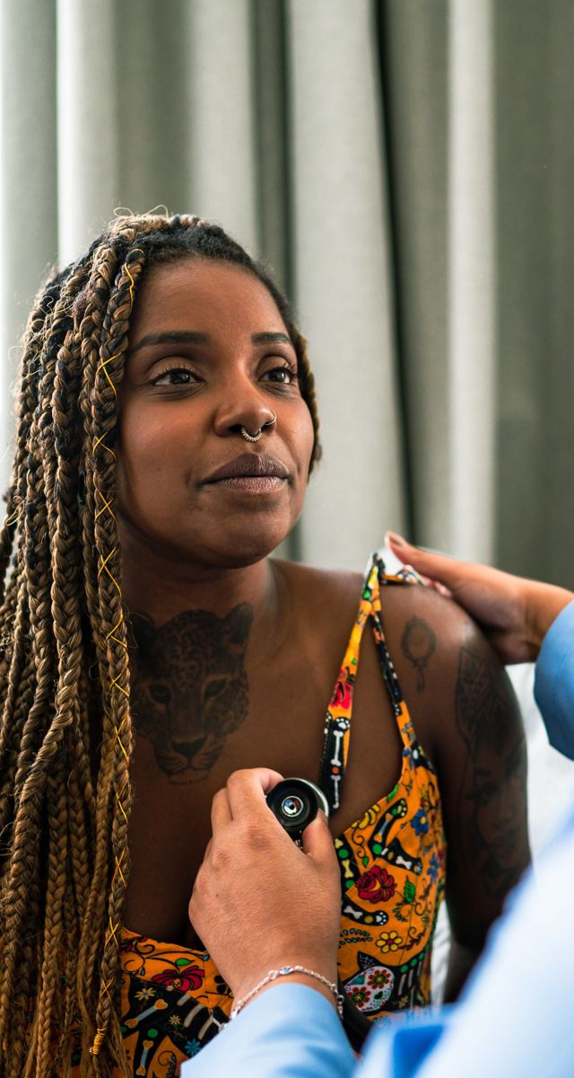 Woman with dark skin and tattoos visiting clinician who is using a stethoscope