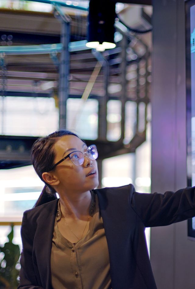 Stock photograph of a young Asian woman conducting a seminar / lecture with the aid of a large screen. The screen is displaying data & designs concerning low carbon electricity production with solar panels & wind turbines. These are juxtaposed with an image of conventional fossil fuel oil production.