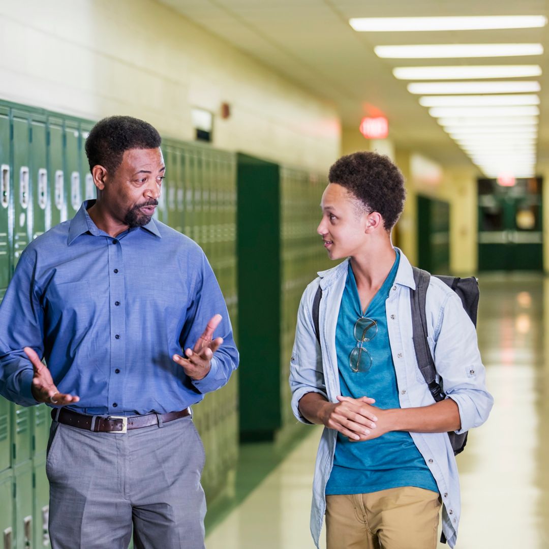 An African-American high school student walking with an adult in the school hallway by a row of lockers. They are talking.