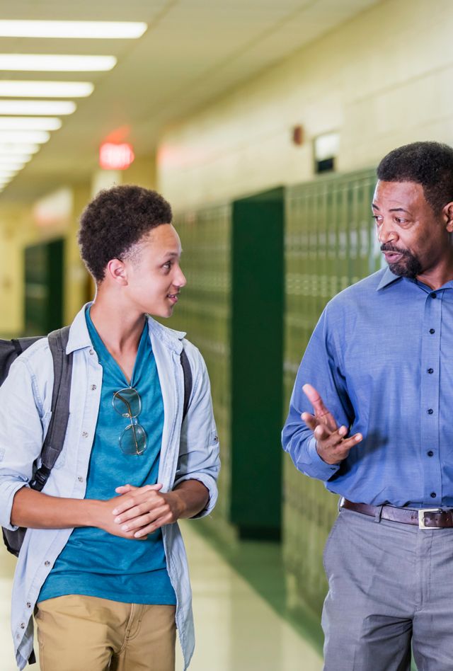 An African-American high school student walking with an adult in the school hallway by a row of lockers. They are talking.