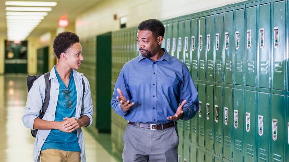An African-American high school student walking with an adult in the school hallway by a row of lockers. They are talking.