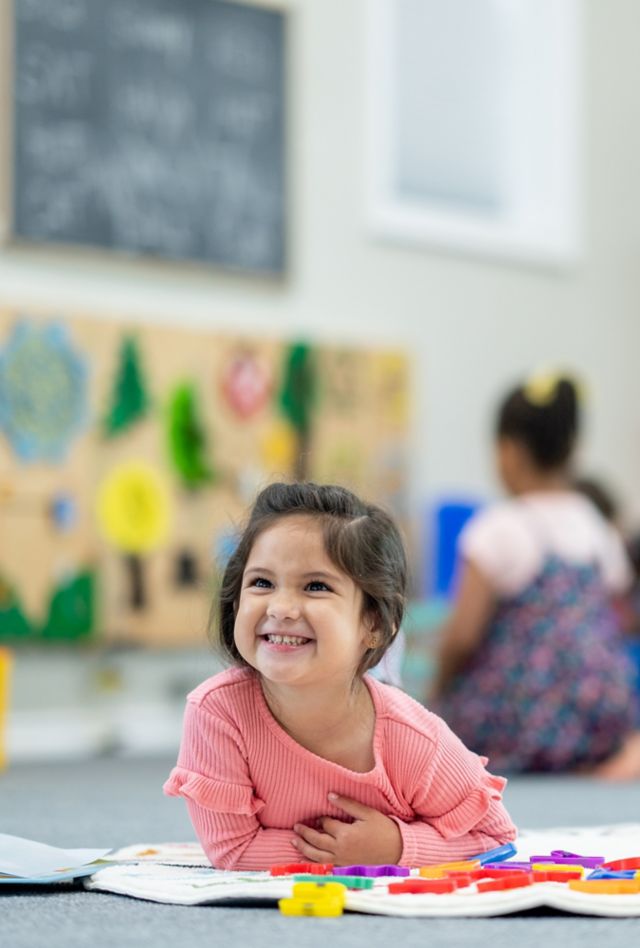 A sweet little girl of Hispanic decent, lays on the rug in her classroom as she plays with an assortment of letters spread out in front of her.  She is dressed casually and smiling.