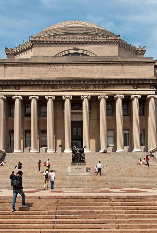 Large stone building with Library of Columbia University written at top, tall ionic columns, and many steps with various people walking and sitting 
