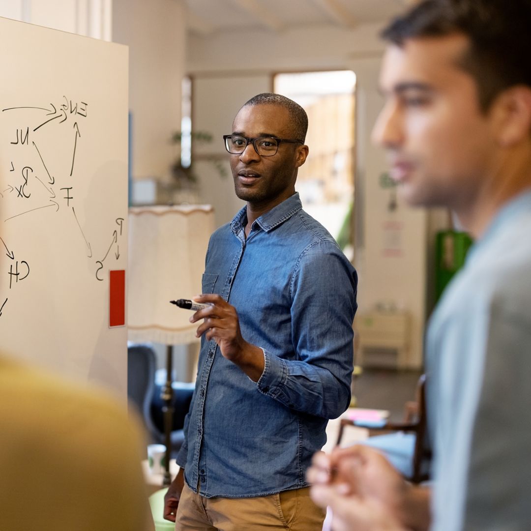Man with dark skin wearing glasses and a denim shirt using a whiteboard to discuss ideas with colleagues at office.