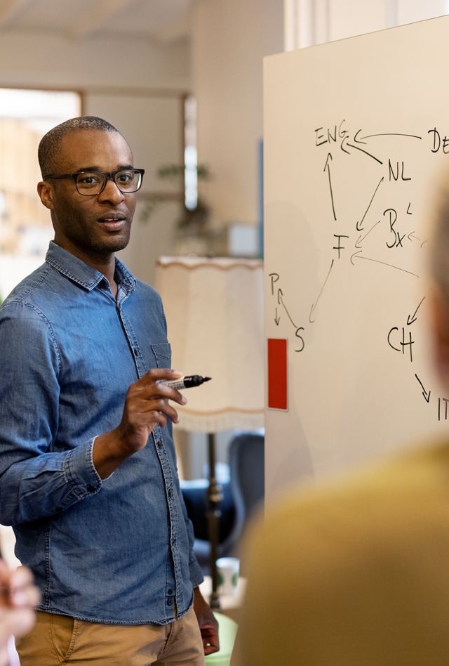 Man with dark skin wearing glasses and a denim shirt using a whiteboard to discuss ideas with colleagues at office. 