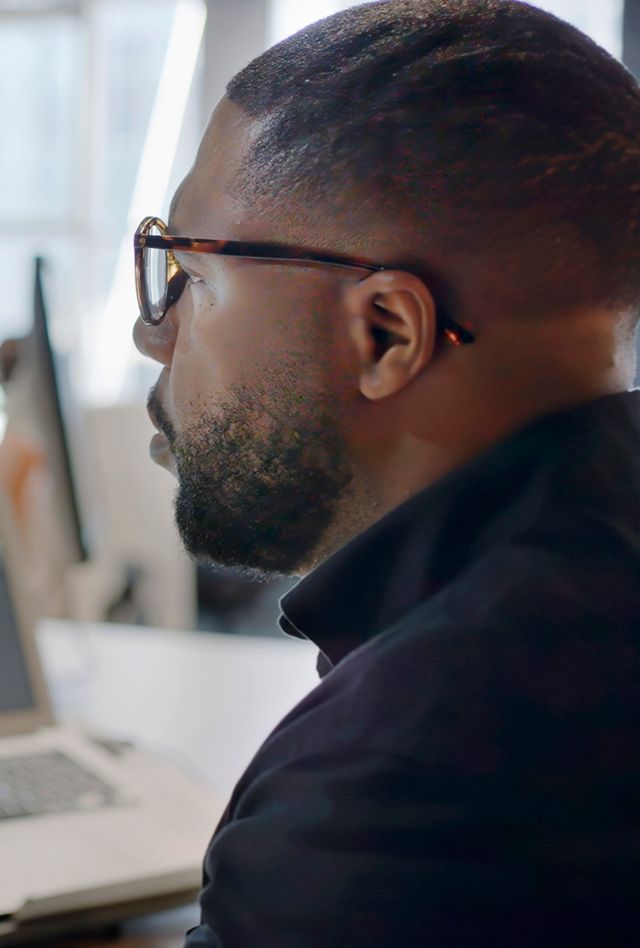 Image showing a Black man’s face looking into a computer screen in an open plan working office. Type is being added to the screen by an Artificial intelligence,  AI,  chatbot.