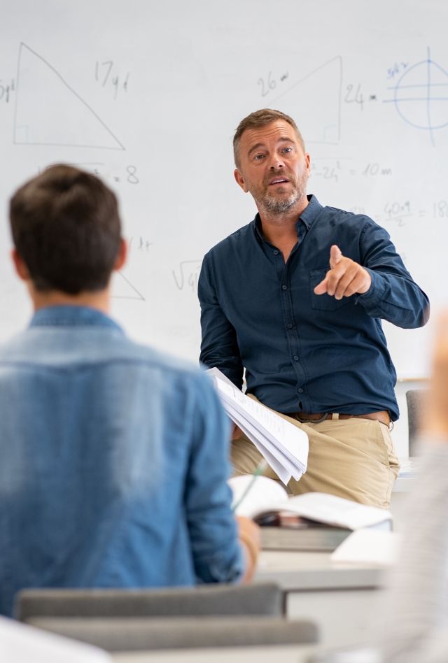 Professor pointing at college student with hand raised in classroom. Student raising a hand with a question for the teacher. Lecturer teaching in class while girl have a question to do during a math lesson.