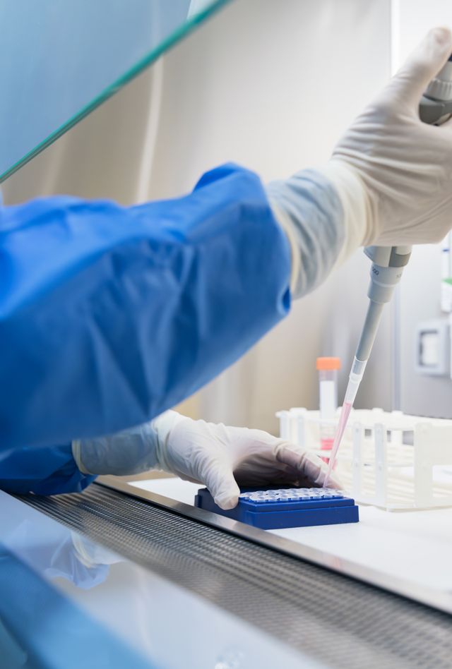 Laboratory setting with a person using a pipette to transfer liquid into a test tube. They are wearing protective gloves and a lab coat. There are test tube racks and lab equipment in the background.