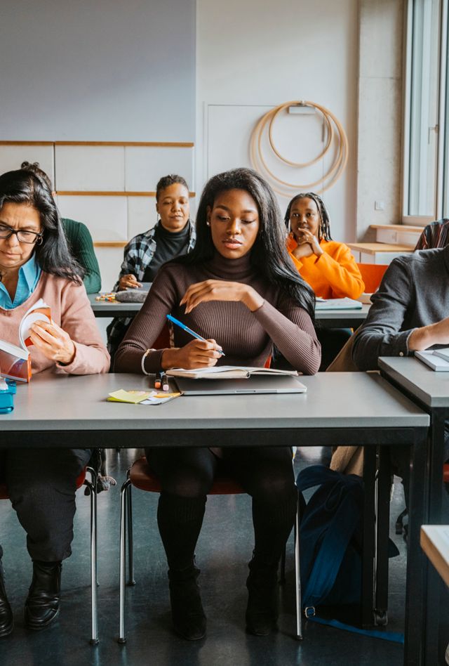 Adult students of various races and sexes sitting at two long tables in a classroom.