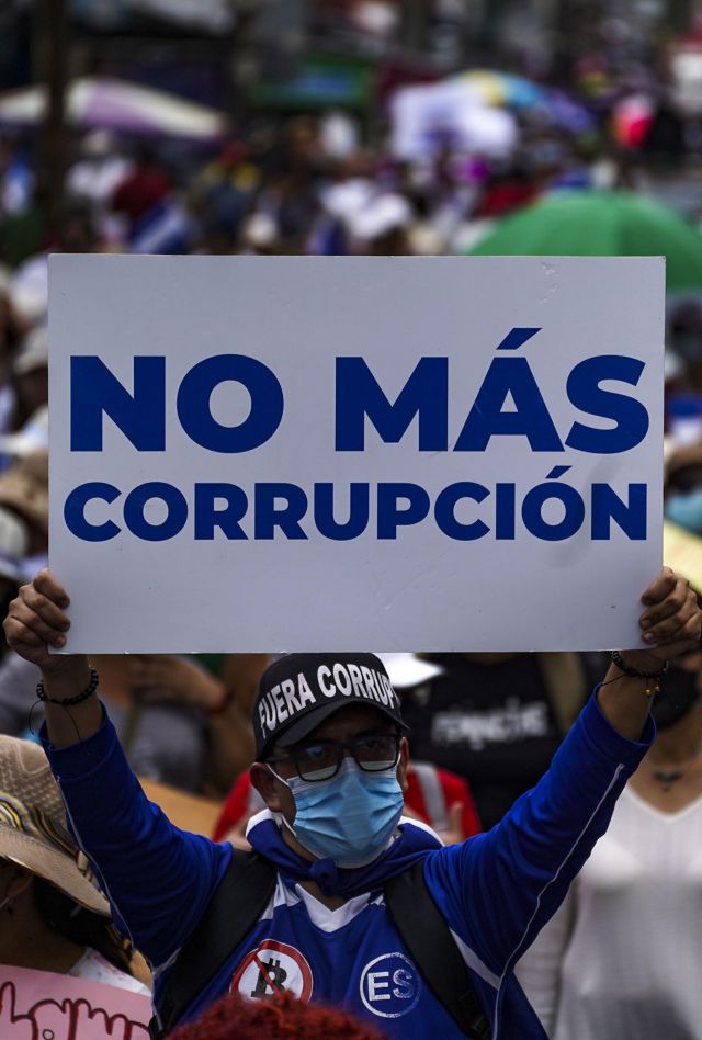 Large crowd marching in the street, with one demonstrator in foreground holding up sign that says "No Más Corrupción"