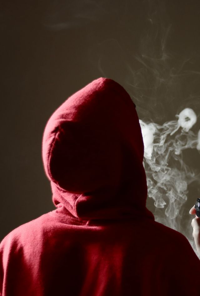 Young male in red hoodie vaping smoking, blows multiple smoke rings while holding a vape in hand, isolated rear view