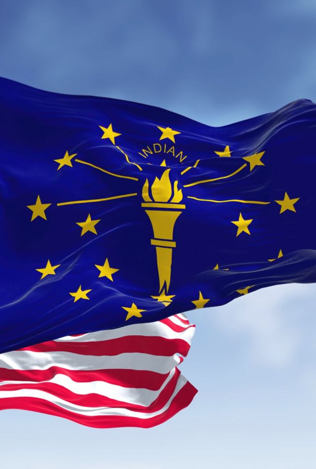 The Indiana state flag waving along with the national flag of the United States of America. In the background there is a clear sky. Indiana is a U.S. state in the Midwestern United States